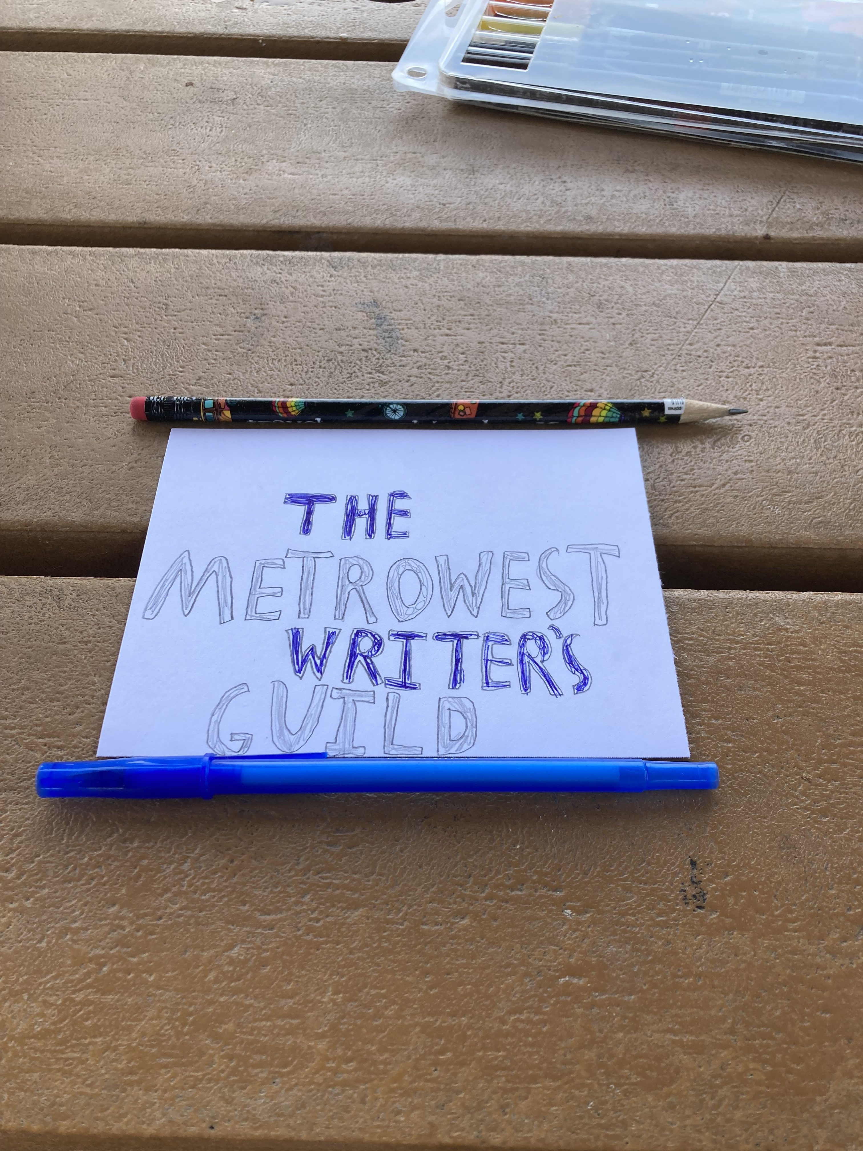 A pen and a pencil along a piece of paper with hand-drawn lettering spelling out "The MetroWest Writer's Guild"